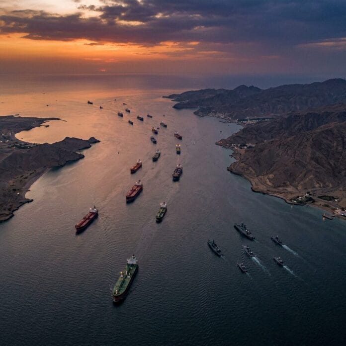 Multiple cargo ships and smaller vessels sailing through a narrow strait with mountainous coastlines at sunset