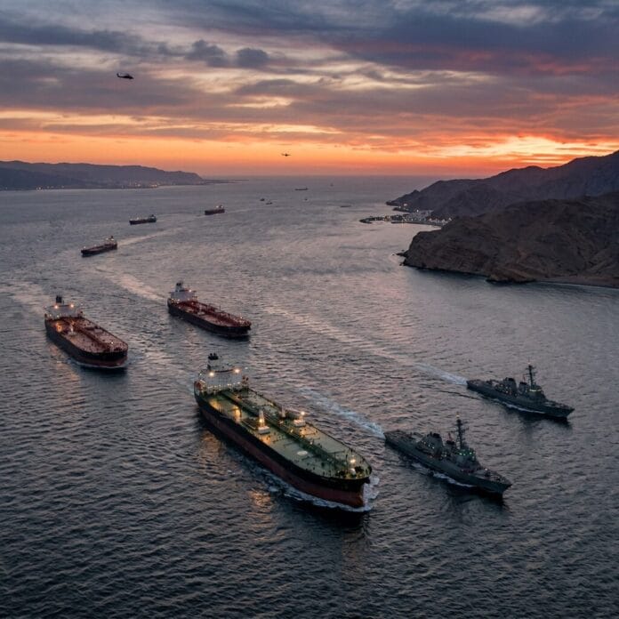 Cargo ships and naval vessels sailing near coastal hills at sunset with helicopters overhead